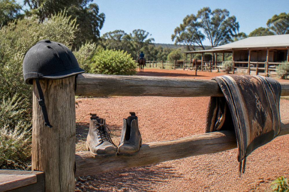A riding helmet and boots resting on a fence rail beside a bush trail